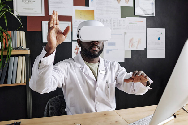 Afrikanischer Arzt mit virtueller Brille gestikuliert in der Luft, während er an seinem Arbeitsplatz im Büro sitzt - Foto, Bild