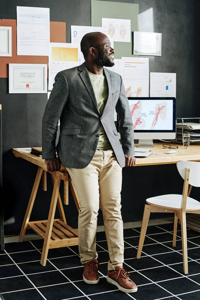 Exitoso médico africano sonriendo y mirando hacia otro lado mientras está de pie en su oficina con computadora y certificados - Foto, Imagen