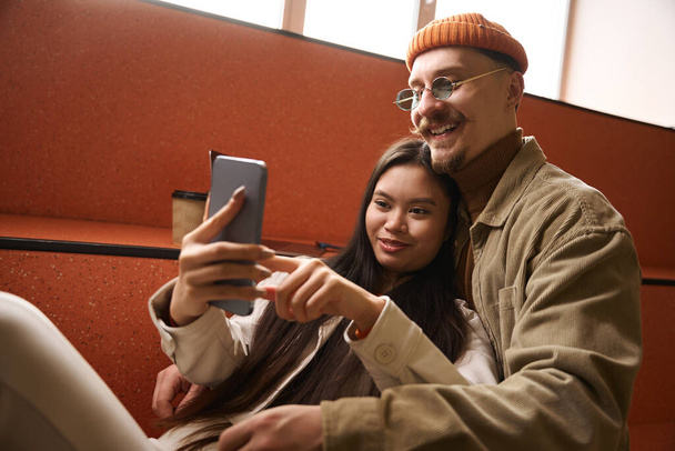 Zufriedene junge Asiatin lächelt und macht Selfie mit fröhlichem Kaukasier - Foto, Bild