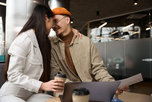 Woman with coffee cup embracing pleased young man seated at laptop with document in hand - Photo, Image