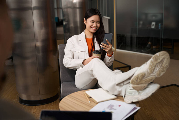 Smiling company employee with paper cup in hands seated in chair with feet on desk looking at smartphone touchscreen - Foto, Bild