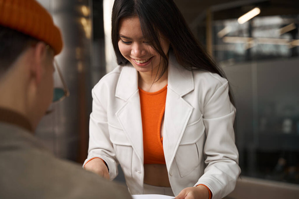 Smiling pleased Asian female office worker showing document to her Caucasian colleague - Foto, imagen