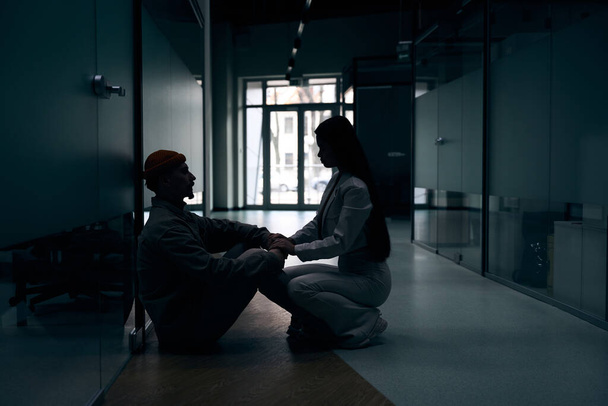 Sympathetic company employee seated on haunches touching hands of her sad colleague in dark corridor - Foto, Imagen