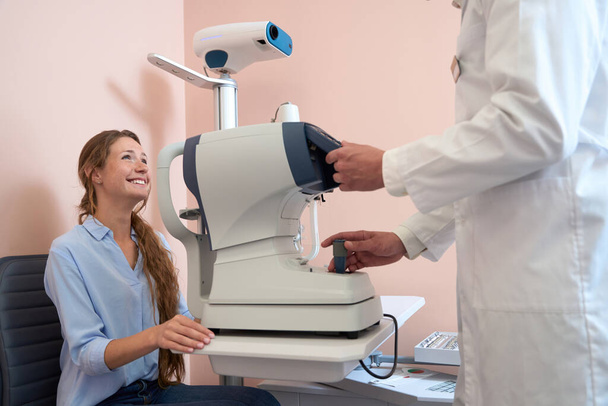 Cintura para cima vista lateral retrato de encantador sorridente paciente feminino está falando com o médico na clínica de oftalmologia - Foto, Imagem