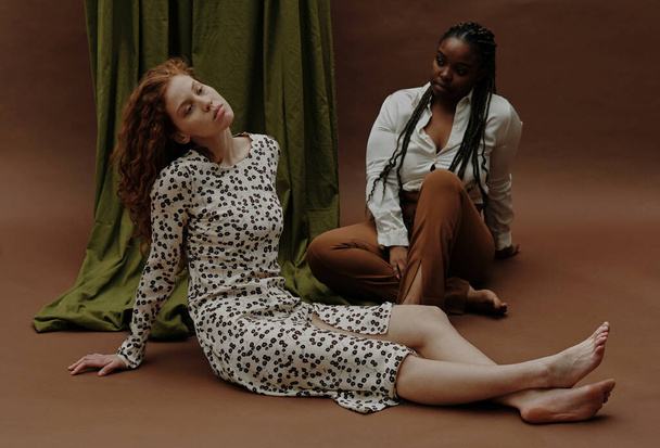 Молодая чувственная белая женщина отдыхает на полу с черной женщиной на заднем плане - Фото, изображение