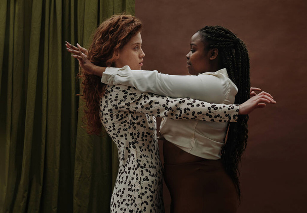 Due ragazze di nazionalità diversa che si abbracciano in piedi sullo sfondo marrone - Foto, immagini