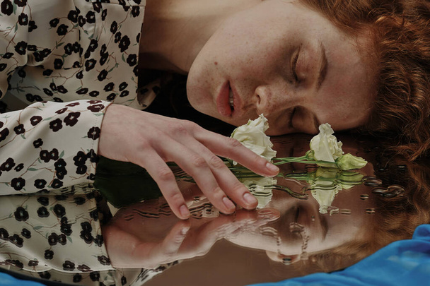 Νεαρή γυναίκα ξαπλωμένη με τα μάτια κλειστά και αγγίζοντας το λουλούδι ξαπλωμένη σε υγρό καθρέφτη - Φωτογραφία, εικόνα