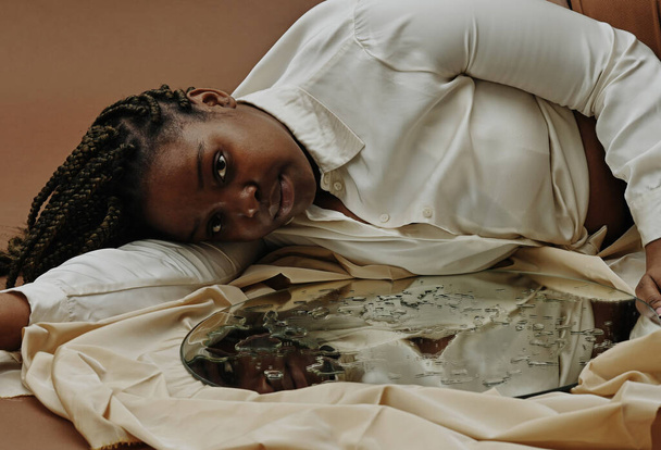 Портрет африканської молодої жінки, що лежить біля дзеркала з краплями води на поверхні і дивиться на камеру. - Фото, зображення