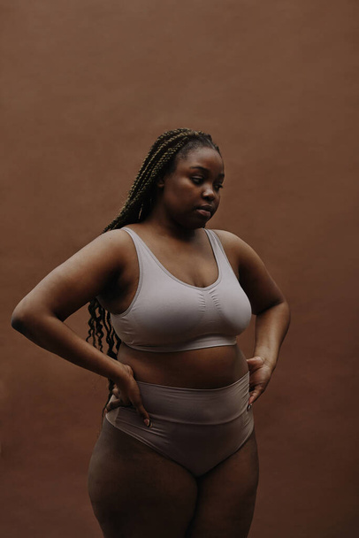 Portrait of African young woman with plump shape standing in underwear against the brown background - Photo, Image