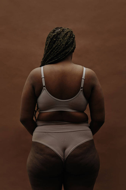Vista posteriore della ragazza africana con figura grassoccia in piedi in biancheria intima contro lo sfondo marrone - Foto, immagini