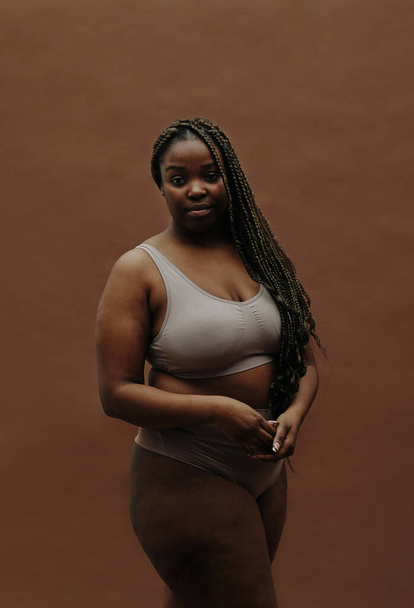 Portrait of African young woman with long braids having plump figure standing in underwear against the brown background - Fotoğraf, Görsel