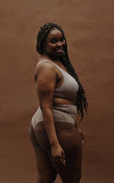 Porträt einer jungen Afrikanerin mit dicker Figur, die isoliert vor braunem Hintergrund in die Kamera lächelt - Foto, Bild
