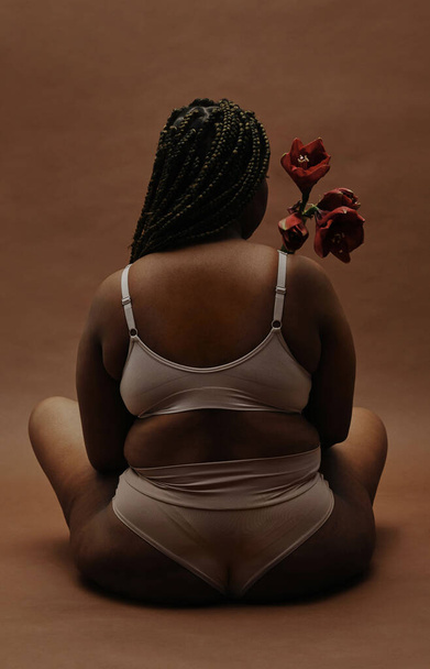 Vue arrière d'une femme africaine en surpoids assise en sous-vêtements et tenant une fleur devant elle isolée sur fond brun - Photo, image