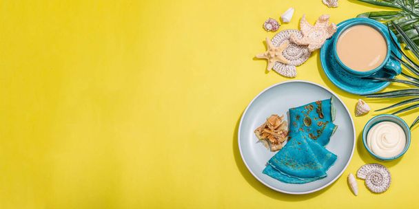 A cup of coffee and blue thin pancakes in a marine style. Starfish, sea shells, palm leaves. Hard light, dark shadow, bright yellow background, banner format - Foto, Imagem
