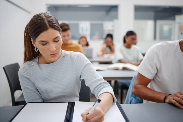 Woman, student and writing university exam in classroom for higher education, scholarship and training. Focused female learning and taking notes in workshop with group of college students. - Photo, Image
