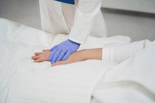 Περικοπή πορτρέτο κεφάλι του μη αναγνωρισμένου γιατρού χέρι σε γάντια αγγίζει το χέρι του ασθενούς, ενώ ο ίδιος βρίσκεται στο ιατρικό κρεβάτι στο νοσοκομείο - Φωτογραφία, εικόνα