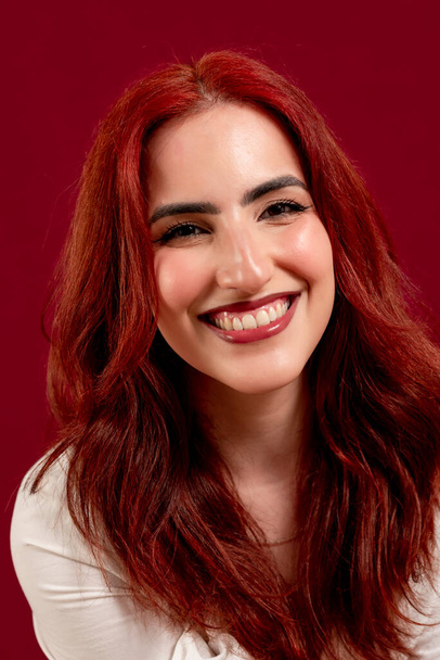Gros plan d'une femme aux cheveux roux confiante regardant la caméra et souriant tout en posant sur un fond rouge isolé. - Photo, image