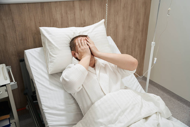 Portrait taille haute d'homme caucasien est couché sur le lit de la médecine avec ses mains sur les yeux dans la salle de médecine - Photo, image