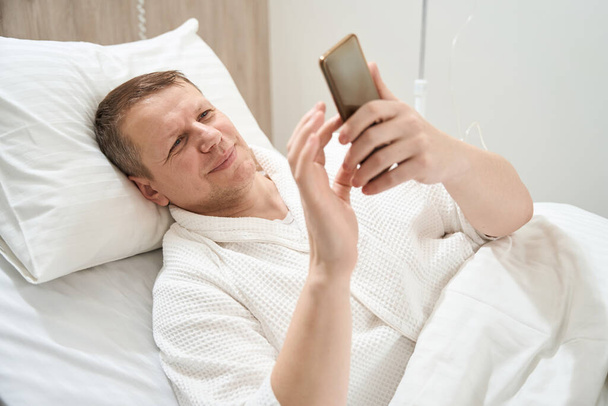 Közelkép portré mosolygó férfi fekszik a gyógyszeres ágyon, miközben néz valamit a mobiltelefon a klinikán - Fotó, kép