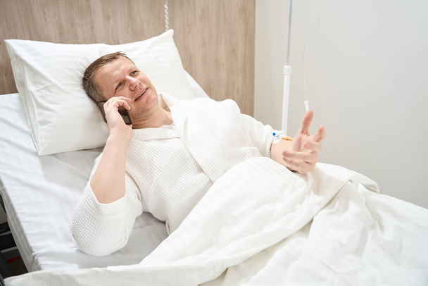 De cerca retrato de vista frontal de hombre valiente sonriente está acostado en la cama médica mientras habla por teléfono móvil en el hospital - Foto, Imagen