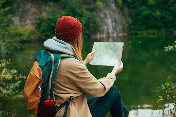 Eine junge Reisende mit Hut sitzt mit einer Papierkarte an einem See und blickt in die Ferne. Berge und See im Hintergrund. Hochwertiges Foto - Foto, Bild