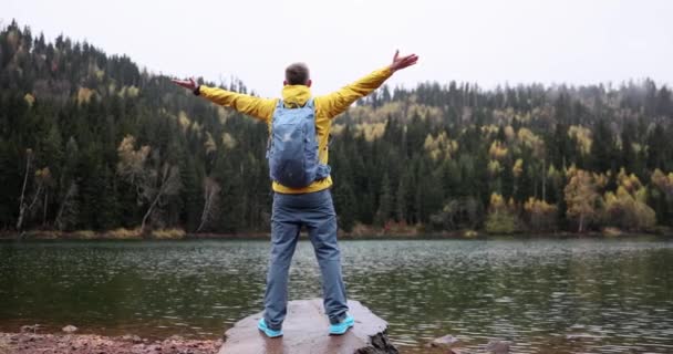 Zadní pohled na mužského turistu s batohem stojící na břehu horského jezera s rukama nad hlavou a s výhledem do údolí. Cestovní dobrodružství a touha - Záběry, video