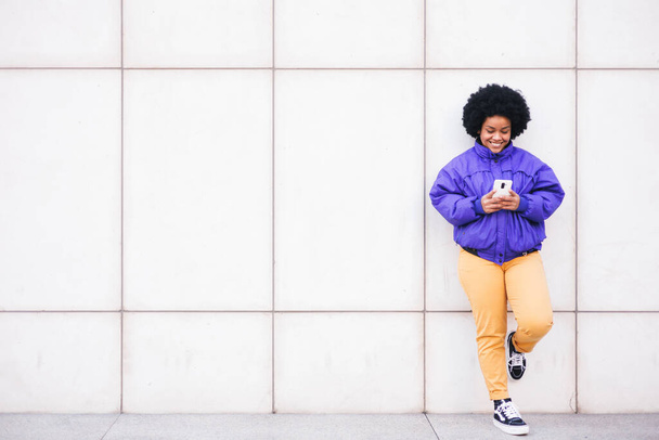 giovane donna afro latina in piedi su parete bianca con telefono e sorridente, spazio per la copia. Foto di alta qualità - Foto, immagini