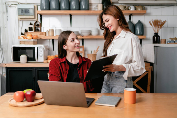 Young woman showing business information on clipboard to friend and reading data together to brainstorming about new project business while working with laptop on the table in kitchen at home. - Фото, зображення