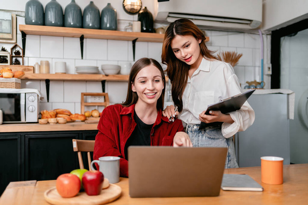 Two young woman pointing data on laptop and reading business information to brainstorming about new project business while working together in the kitchen at home. - Photo, image