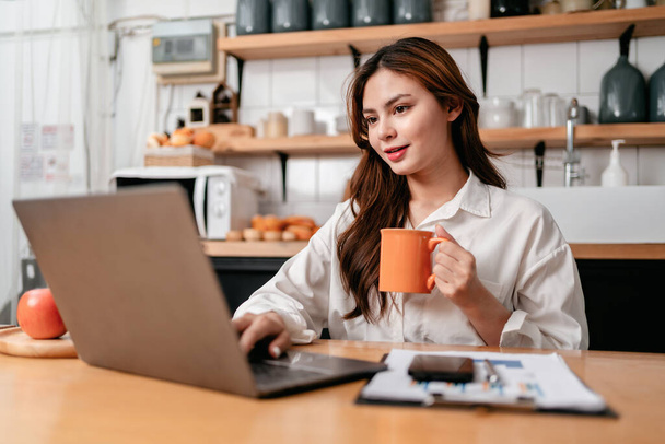 Jonge zakenvrouw drinken van koffie en typen om het zoeken van zakelijke informatie op de laptop, terwijl zitten te werken en denken strategie over nieuwe business project in de keuken thuis. - Foto, afbeelding