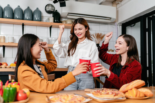 Grupo de amigos tomando un vaso de bebida y comiendo pizza italiana con pan juntos mientras disfrutan y levantan el brazo para celebrar en la fiesta en la cocina en casa. - Foto, imagen