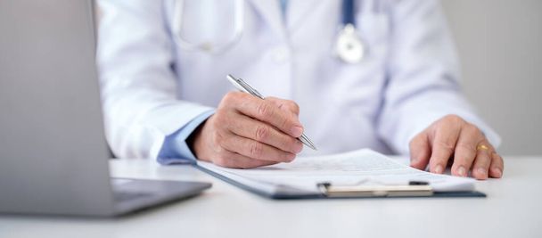 Senior mannelijke arts met stethoscoop diagnose van de ziekte van de patiënt en het schrijven van recept op klembord, terwijl het werken aan het zoeken naar behandeling van de ziekte informatie op laptop in het ziekenhuis. - Foto, afbeelding