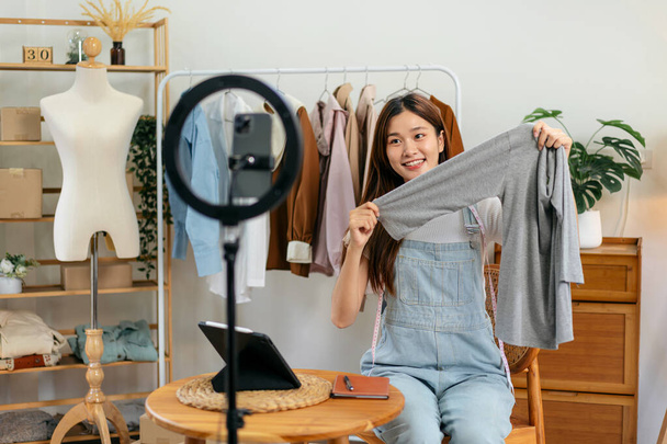 Γυναίκα online πωλητής χρησιμοποιεί smartphone για να μεταδώσει ζωντανά στο Vlog για την εισαγωγή νέων μόδας, ενώ αναθεώρηση συλλογή πουκάμισο και μιλώντας με τον πελάτη για την πώληση online ρούχα στα μέσα κοινωνικής δικτύωσης. - Φωτογραφία, εικόνα