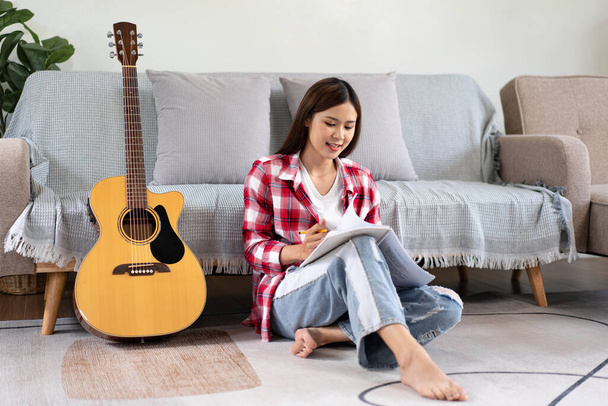 Giovane donna è comporre la canzone e la scrittura di testi su notebook dopo aver suonato la chitarra mentre seduto sul pavimento in soggiorno a casa. - Foto, immagini