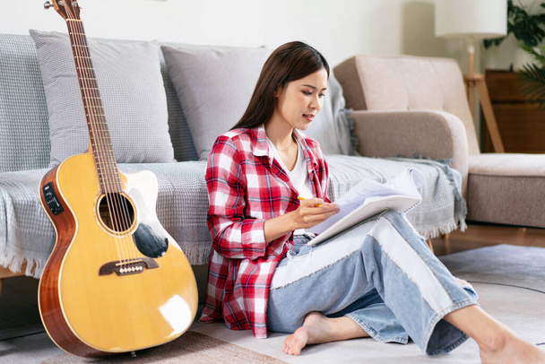 Mujer joven es componer la canción y escribir letras en el cuaderno después de tocar la guitarra mientras está sentado en el suelo en la sala de estar en casa. - Foto, imagen