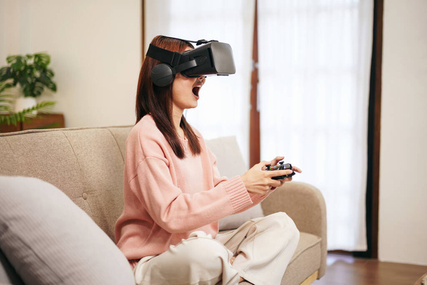 Evde oturma odasında dinlenirken elinde joystick ile sanal gerçeklikle video oyunu oynayan süveter ve sanal gözlük takan genç bir lezbiyen kadın.. - Fotoğraf, Görsel