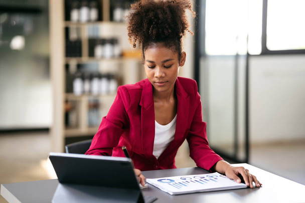 African american businesswoman in suit reading accounting information of business on tablet and paperwork to taking note in notebook while working and thinking strategy of project in modern workspace. - Zdjęcie, obraz