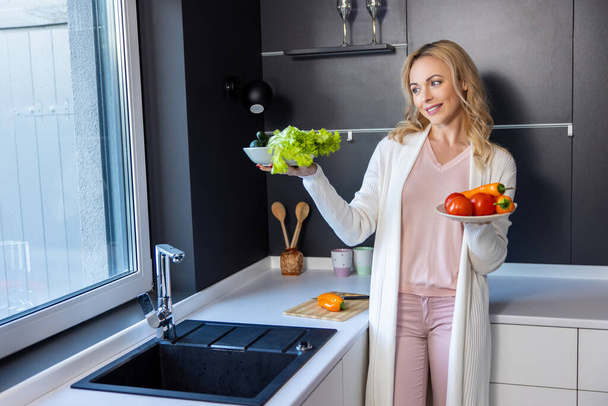 Joyous young female leaning against the kitchen counter while holding two plates with fresh vegetables - Fotoğraf, Görsel