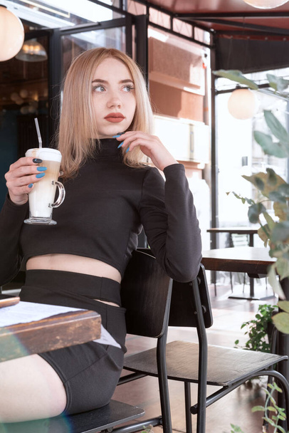 Portrait of young blonde woman in cafe. Girl enjoys latte coffee in the cafeteria. Vertical frame - Foto, afbeelding