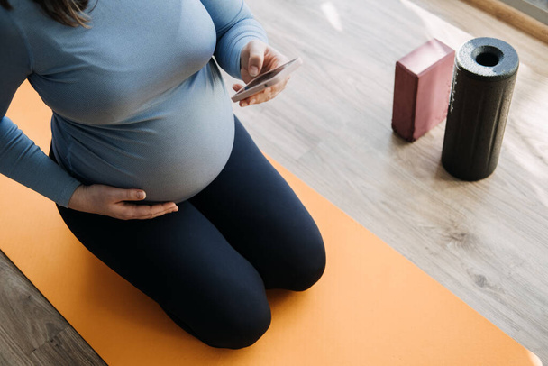 Hamilelik Egzersizleri, Hamilelik Uygulamaları. Doğum Öncesi ve Doğum Sonrası Antrenmanı, Hamilelik Sağlığı ve Yoga Çalışmaları cep telefonunda. Hamile kadın telefonda antrenman yapmak istiyor.. - Fotoğraf, Görsel