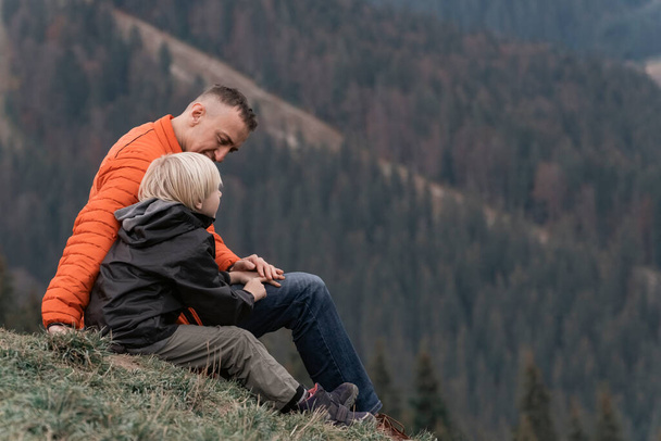 O pai e o filho sentam-se na encosta e olham para as montanhas. Trekking familiar com crianças na floresta. - Foto, Imagem
