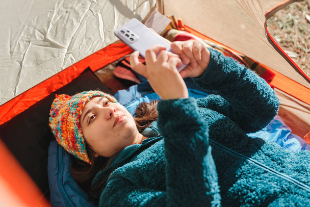 若い旅行者の女性は暖かい服を着て、彼女の携帯電話を使用して、午前中にキャンプテントの中に横たわっていた。高品質の写真 - 写真・画像