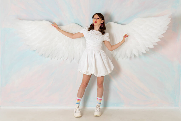 Симпатична дівчина-ангел брюнетка у вишуканій короткій білій сукні, величезних крилах пір'я та яскравому макіяжі на фоні неба
. - Фото, зображення