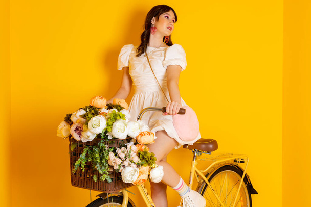 Cute stylish brunette girl, dressed in a fancy short white dress and bright makeup is sitting on a bike with flowers and happily posing and looking away. Isolated on yellow. - Photo, Image