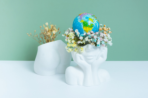 Earth day, save the planet concept. Plaster vase head-shape with flowers and world globe and ass with dry flowers. - Фото, изображение
