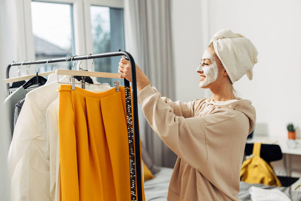 A happy Caucasian woman with a mask on her face is choosing clothes to wear - Фото, зображення