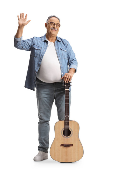 Full length portrait of a mature man standing with an acoustic guitar and waving isolated on white background - Foto, imagen