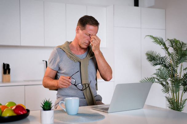 Fatigued mature old man taking off glasses suffering from tired dry irritated eyes after long computer use, senior middle aged male feels eye strain problem or blurry vision working on laptop at home - 写真・画像