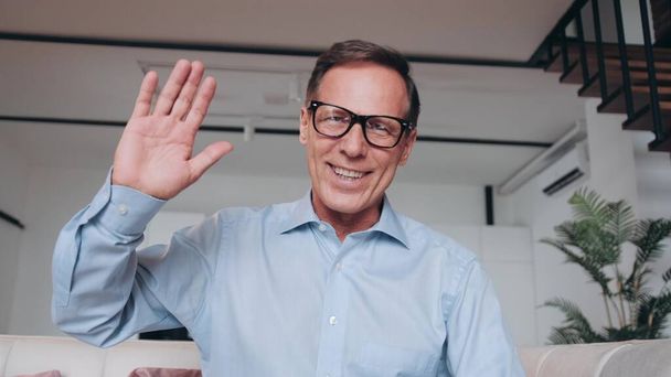 Happy mature 60s man in glasses waving hello at camera, smiling, laughing, speaking on video call at home. - Foto, imagen