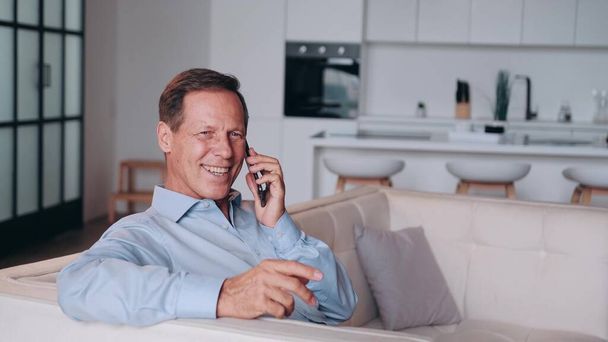 Successful and smiling middle-aged man in blue shirt talking by the phone at home. Sitting on the couch. Living room interior is blurred in background - Photo, image
